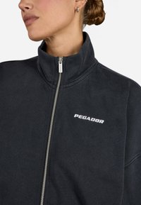 Juoda zip-up striukė iš minkštos medžiagos su aukštu apykakle. Turėti sidabrinę užtrauktuką ir baltą "PEGADOR" logotipą kairėje krūtinės pusėje.