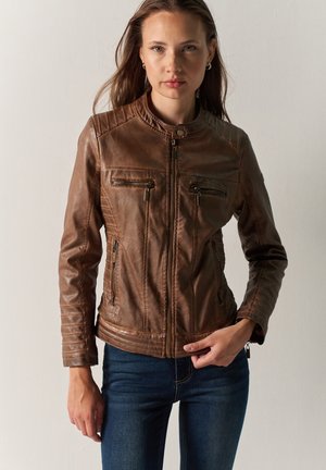 Femme portant une veste en cuir marron ajustée avec des poches zippées et un jean bleu, debout devant un fond clair uni.