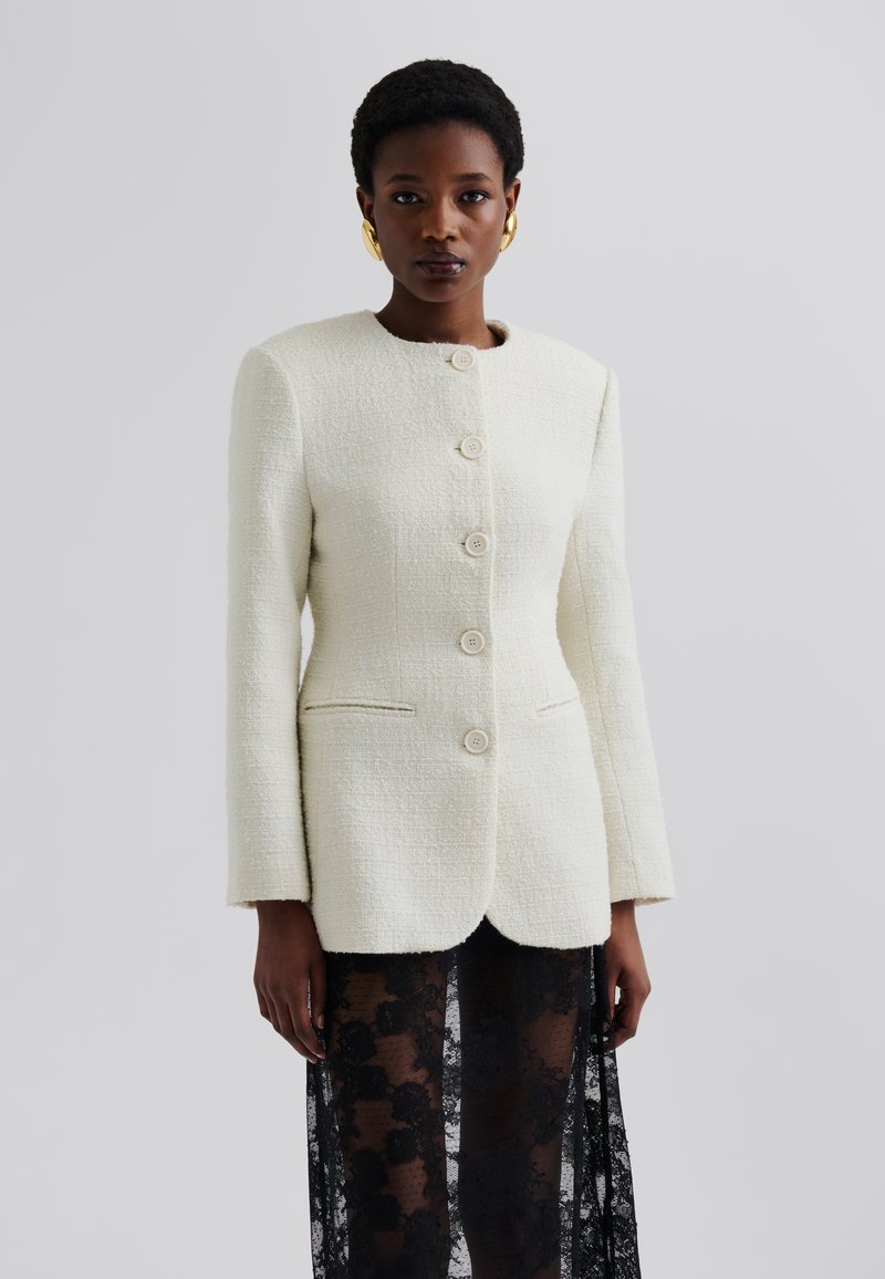Femme portant une veste blanche texturée boutonnée et une jupe en dentelle noire transparente, accessoirisée de grandes boucles d'oreilles créoles dorées, se tenant devant un fond gris.