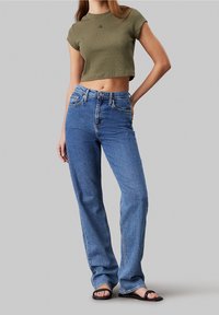 Zkrácený olivově zelený tričko s malým logem, v kombinaci s modrými džíny s vysokým pasem. Outfit je doplněn černými sandály.