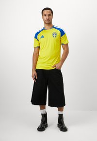 Man standing wearing yellow and blue Sweden soccer jersey, black wide shorts, white socks, and black ankle boots against a plain background.