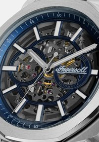 Syrgastålsklocka med en blå bezel, transparent urtavla som visar detaljerade kugghjul, silver- och guldkänsla med tydliga timmarkörer.