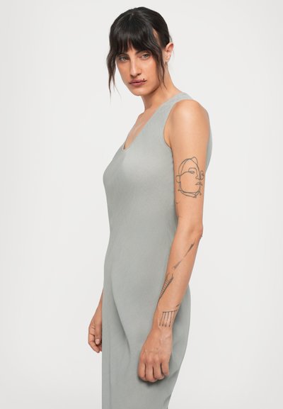 Woman with dark hair and bangs wearing a sleeveless gray dress, showing abstract tattoos on her left arm against a plain white background.