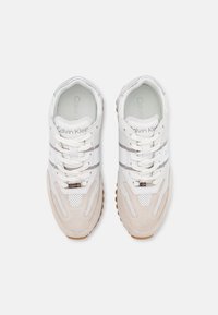 Sneaker atletiche bianche con pannelli in rete e camoscio, suola in gomma testurizzata e branding prominente. Presentano un design con lacci e accenti sottili.