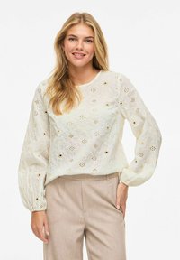 Femme blonde souriante, portant un chemisier blanc à manches longues brodé et un pantalon beige taille haute, devant un fond uni.