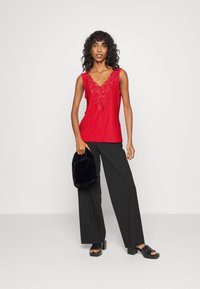 Top sem mangas vermelho com detalhe em bordado no decote em V, combinado com calças largas pretas e uma bolsa de pelúcia negra. Sandálias de salto bloco pretas.