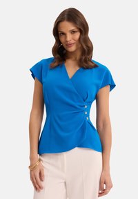 Niebieska bluzka z krótkimi rękawami w stylu kapslowym, z teksturowanej tkaniny, z efektownym przodem i złotymi guzikami z boku, w zestawieniu z jasnymi spodniami.