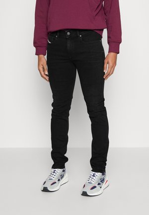 Person trägt schwarze Slim-Fit-Jeans, weiße und marineblaue Sneaker und ein dunkelrotes Langarmshirt, steht vor einem einfarbigen hellen Hintergrund.