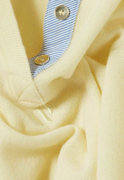 Jersey amarillo de punto con una textura suave, que presenta un cuello a rayas azul y blanco y botones dorados, mostrando un aspecto arrugado.