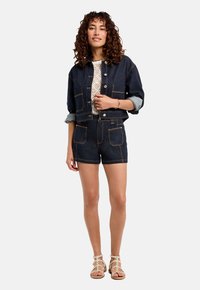 Veste en denim noir à manches retroussées, fermeture à boutons et poches avant, accompagnée d'un short en denim assorti et de sandales à lanières.