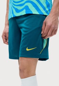 Άνδρας που φοράει τιρκουάζ αθλητικές σορτς Nike με κίτρινο λογότυπο και πράσινο πουκάμισο με σχέδιο, στέκεται με ορατές άσπρες κάλτσες σε ανοιχτό φόντο.