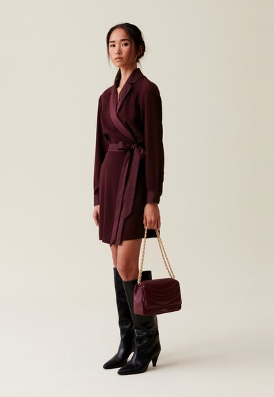 Vestido envolvente burdeos con mangas largas y cinturón de lazo, combinado con botas negras hasta la rodilla y un bolso burdeos con asa de cadena.
