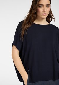 Jonge vrouw met lang bruin haar, gekleed in een loszittend donkerblauw kortärmelig shirt, met hand in de zak, kijkt recht vooruit tegen een lichte achtergrond.