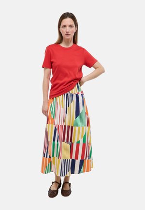 Femme debout, main sur la hanche, portant un t-shirt rouge à manches courtes, une jupe à motifs géométriques colorés et des chaussures sombres tissées.