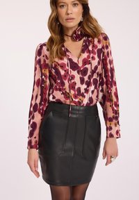 Kobieta ubrana w bluzkę z długim rękawem w różowo-fioletowy wzór z kwiatowym detalem przy szyi oraz czarną skórzaną spódnicę z kieszeniami.