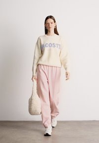 Λευκό φλις πουλόβερ με το λογότυπο "Lacoste", συνδυασμένο με ροζ tapered joggers. Το μοντέλο κρατάει μια υφασμάτινη μπεζ τσάντα και φοράει λευκά αθλητικά παπούτσια.
