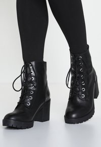 Botas de tobillo de cuero negro con un tacón alto y grueso, parte frontal con cordones y ojales de metal; suela robusta con un dibujo para un mejor agarre.