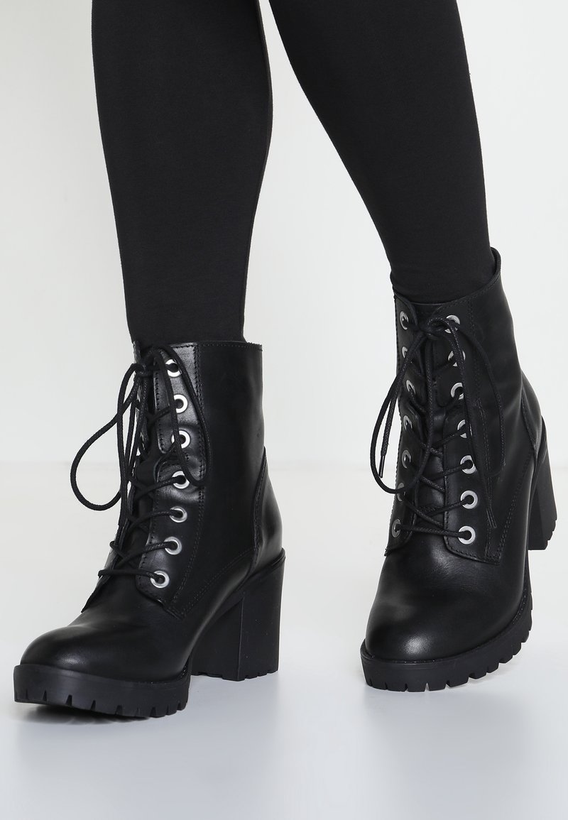 Botas de tobillo de cuero negro con un tacón alto y grueso, parte frontal con cordones y ojales de metal; suela robusta con un dibujo para un mejor agarre.