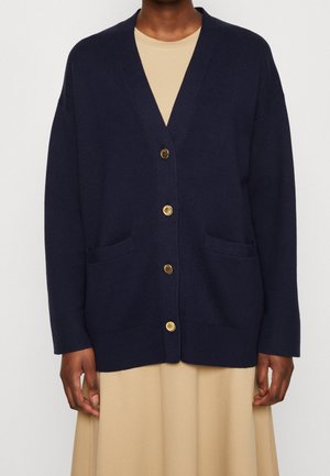 Osoba ubrana w granatowy kardigan zapinany na guziki z złotymi guzikami, na beżowej sukience, pokazana z przodu od szyi do połowy uda.