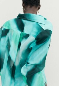Une chemise légère et transparente turquoise avec un motif dégradé, dotée d'un col classique et de manches longues, présentant des variations texturales fluides.