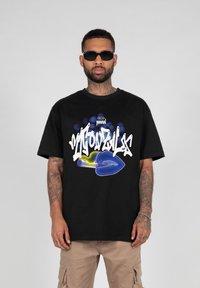 Sort t-shirt med fed hvid og blå grafisk design, der viser læber og en mikrofon. Bæres med brune cargo bukser og mørke solbriller.