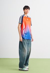 Oranžinės ir mėlynos gradientinės spalvos sportinė marškinėliai su baltais akcentais, derinami su laisvomis pilkomis džinsinėmis kelnėmis ir žaliai dryžuotais baltais sportbačiais.
