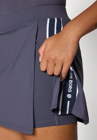 Pelēka sporta svārki ar krokām, ar zilu un balto svītru detaļām. Roka tur iekšējo kabatu ar zīmolu. Gluda tekstūra.