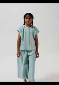 Pige med fletninger iført lyseblå blomsterprintet skjorte med knapper og matchende vide bukser, stående foran en ensfarvet hvid baggrund.