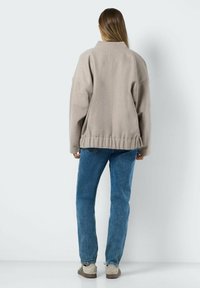 Veste en polaire beige surdimensionnée avec un col montant et un ourlet élastique, associée à un jean en denim bleu clair et des chaussures beiges, vue de dos.