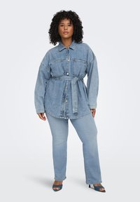 Γυναίκα με σγουρά μαλλιά που φοράει φαρδιά τζιν μπουφάν με ζώνη, ανοιχτόχρωμα μπλε τζιν και πεδιλάκια σε τιρκουάζ χρώμα, στέκεται μπροστά σε απλό φόντο.