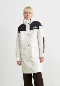 Veste imperméable blanche avec des accents noirs, col haut, capuche ajustable, boutons à l'avant et deux poches avant. Logo sur la poitrine.