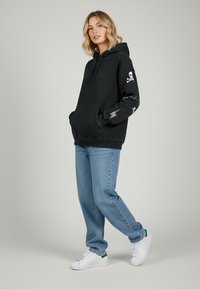 Felpa nera con cappuccio, decorata con grafiche di teschio e ossa incrociate, abbinata a jeans azzurri chiari e sneakers bianche con dettagli verdi.