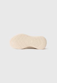 Beige skosula med intrikata zigzag- och vågmönster, med en upphöjd tumme-upp-symbol i mitten.