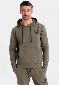 Sudadera color verde olivo de tejido suave, con un bolsillo tipo canguro, cordones negros y un parche cuadrado en el pecho. Corte casual.