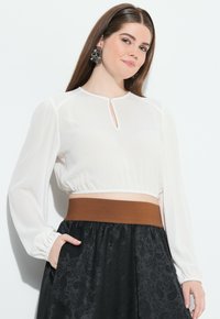 Weißes, kurzes Blusenshirt mit Puffärmeln, das mit einem geknöpften Schlüsselloch-Ausschnitt am Hals versehen ist. Kombiniert mit einem schwarzen Rock mit strukturierten Akzenten.