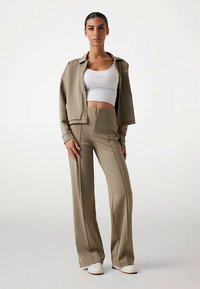 Giacca corta color cammello e pantaloni a gamba larga abbinati in tessuto liscio, abbinati a un crop top bianco e sneakers bianche.