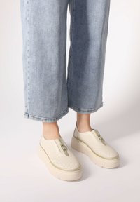Béžové perforované slip-on boty s metalickým akcentem na zipovém uzávěru, se silnou platformovou podrážkou, v kombinaci s světlými modrými širokými džíny.