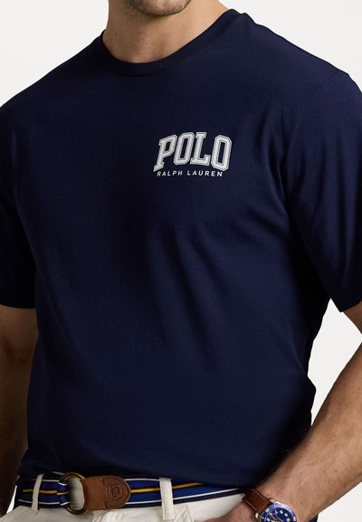 Tumši zils kokvilnas T-krekls ar baltajiem "POLO" un "RALPH LAUREN" logotipiem, apaļa kakla izgriezums, īsās piedurknes, valkāts kopā ar krāsainu jostu.