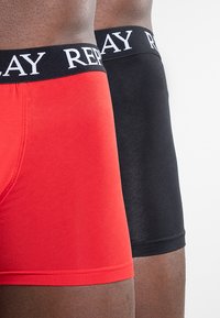 Dos pares de bóxers, uno rojo y otro negro, con cinturillas elásticas que tienen "REPLAY" impreso en blanco. Textura de tela suave.