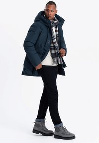 Mörkblå puffjacka med huva, vit tröja, rutmönstrad halsduk, mörka jeans och grå stövlar. Jackan har sidofickor och dragkedjestängning.