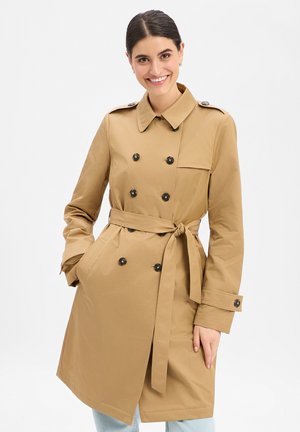 Smiling woman with dark hair wears a knee-length tan trench coat, tied at the waist, with double-breasted buttons and hands in pockets.