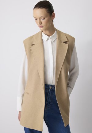 Beige liivita blazer, kus on kohandatud krae, kaks esist pocketit, kantud valge särgi kohal, koos siniste kõrge vöökohaga teksadega.