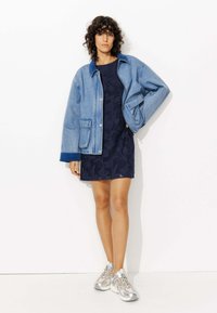 Veste en denim avec une coupe décontractée, associée à une robe en dentelle marine et des chaussures de sport argentées. Comprend une fermeture à boutons et de grandes poches.