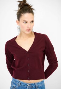 Cardigan corto bordeaux con scollatura a V e cinque bottoni. Maniche lunghe, texture morbida e design aderente. Abbinato a pantaloni corti in denim a vita alta.