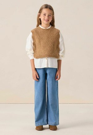 Meisje van jonge leeftijd met een glimlach, gekleed in een bruine gebreide vest over een witte longsleeve, wijde blauwe jeans en bruine schoenen, staand tegen een effen achtergrond.