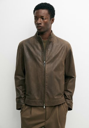 Man wearing a brown zip-up leather jacket and matching brown pants, standing with hands in pockets, looking to the side against a white background.