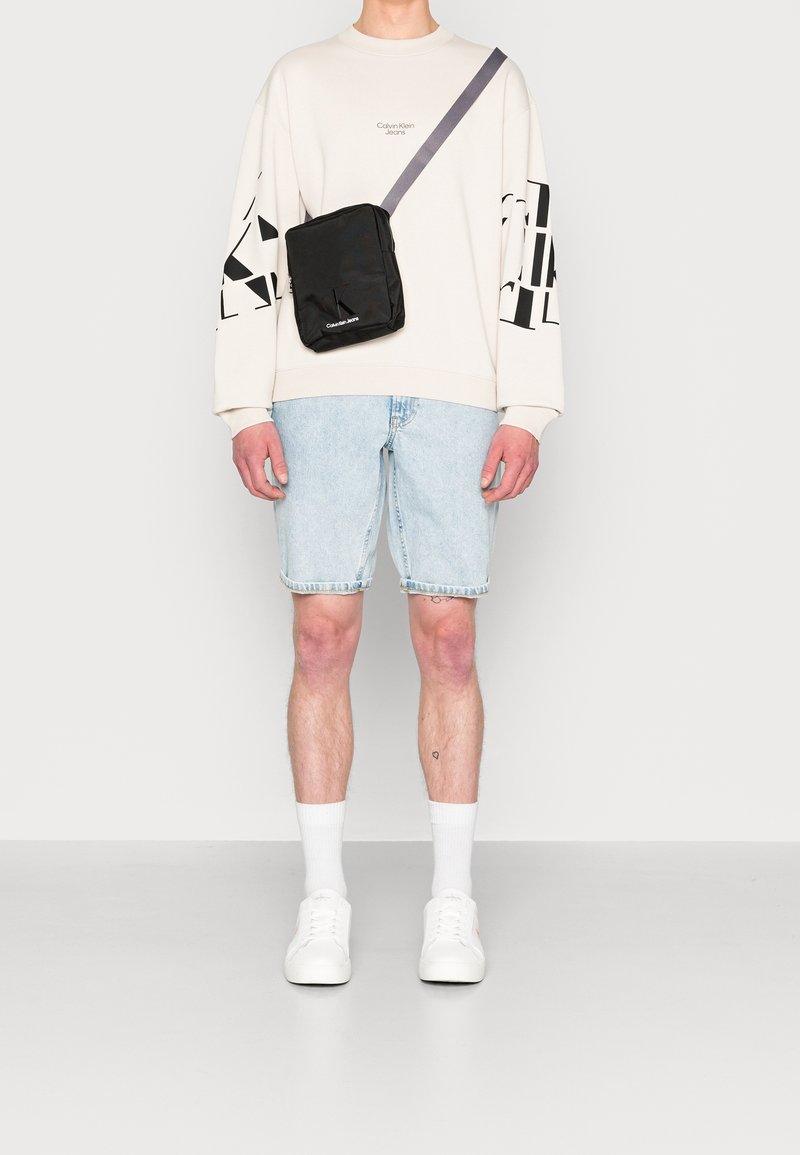 Personne portant un sweatshirt blanc Calvin Klein, un short en denim bleu clair, des chaussettes blanches, des baskets blanches et un sac en bandoulière noir.