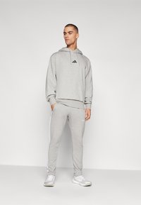 Sweatshirt atlético cinza com um logotipo preto, combinado com calças de treino cinzas a condizer, apresentando riscas brancas laterais e um design ajustado.