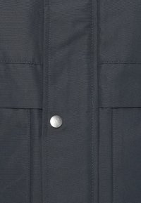 Tessuto grigio scuro e strutturato con chiusura anteriore a scatto e due tasche sul petto con dettaglio cucito per maggiore durata.
