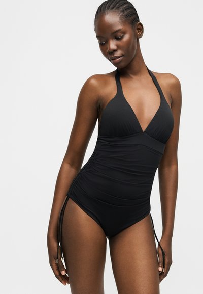 Γυναίκα με πλεξούδες στα μαλλιά που φοράει μαύρο ολόσωμο μαγιό με λαιμόκοψη halter, ραφές στο πλάι και κορδόνια, στέκεται μπροστά σε απλό ανοιχτό φόντο.